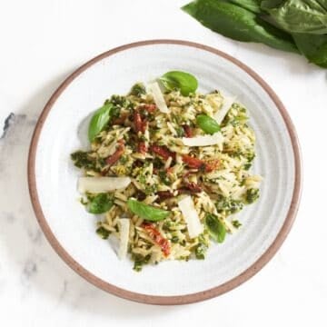 A plate of orzo pesto pasta salad with sun dried tomatoes topped with grated parmesan cheese and fresh basil leaves, a bunch of fresh basil leaves are in the upper right.