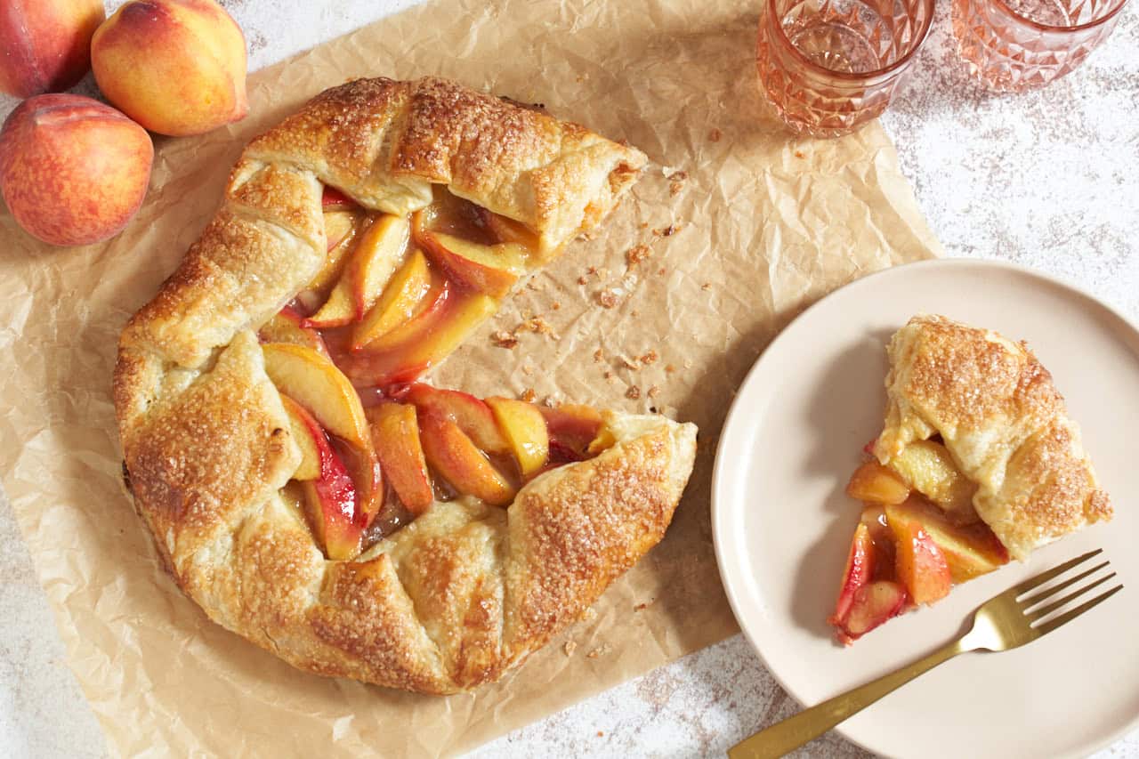 A peach puff pastry galette with a slice cut out of it on parchment paper, a pink plate with a slice of galette is on the right, two pink glasses and three peaches are in the upper corners.