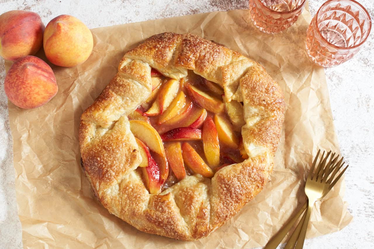A peach puff pastry galette on parchment paper, three gold forks are on the right, two pink glasses and three peaches are in the upper corners.