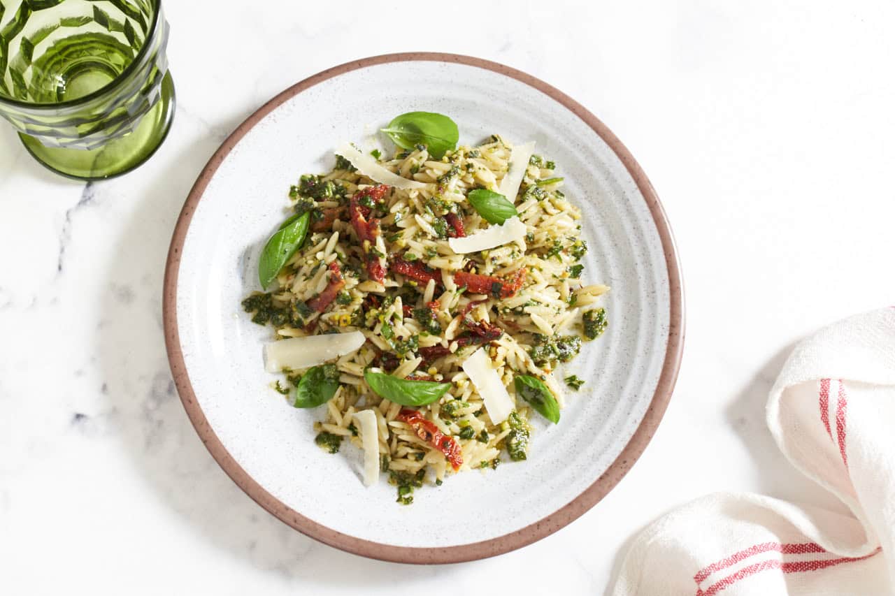 A plate of orzo pesto pasta salad with sun dried tomatoes topped with grated parmesan cheese and fresh basil leaves, a green glass is in the upper left, a red and white striped towel is in the bottom right.