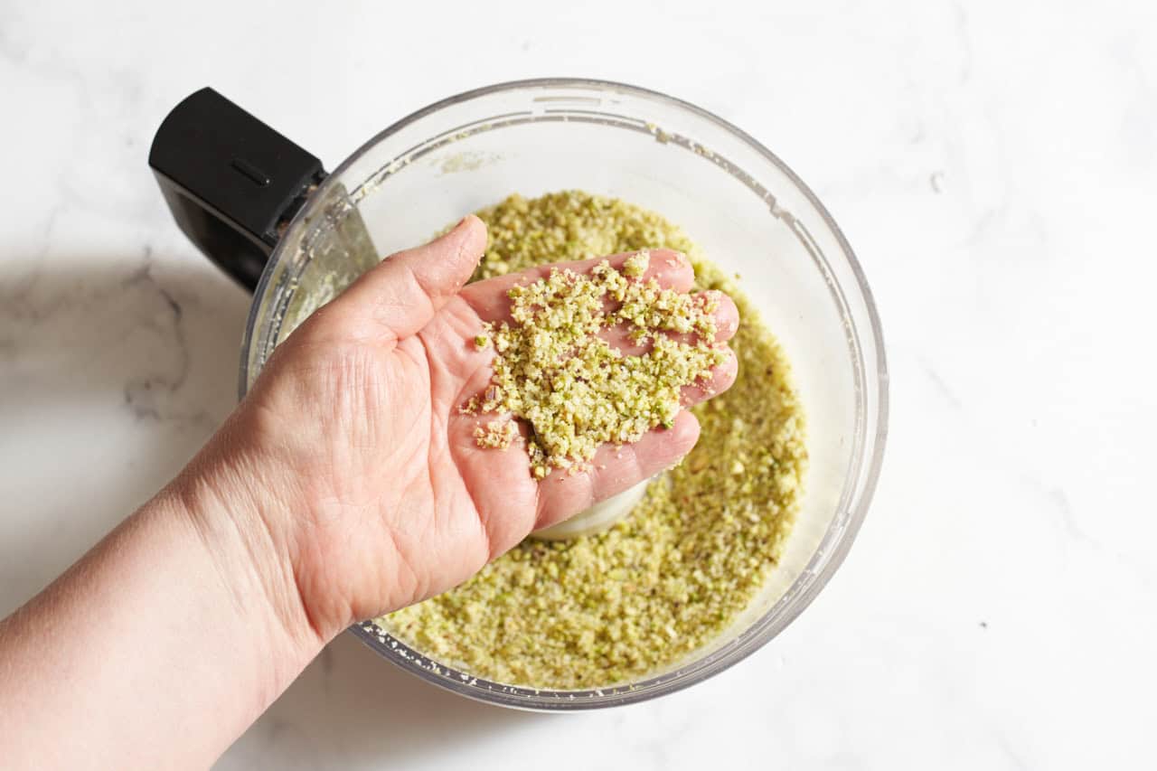 A woman's hand over a food processor holding parmesan and pistachios that have been pulsed into a coarse mixture.