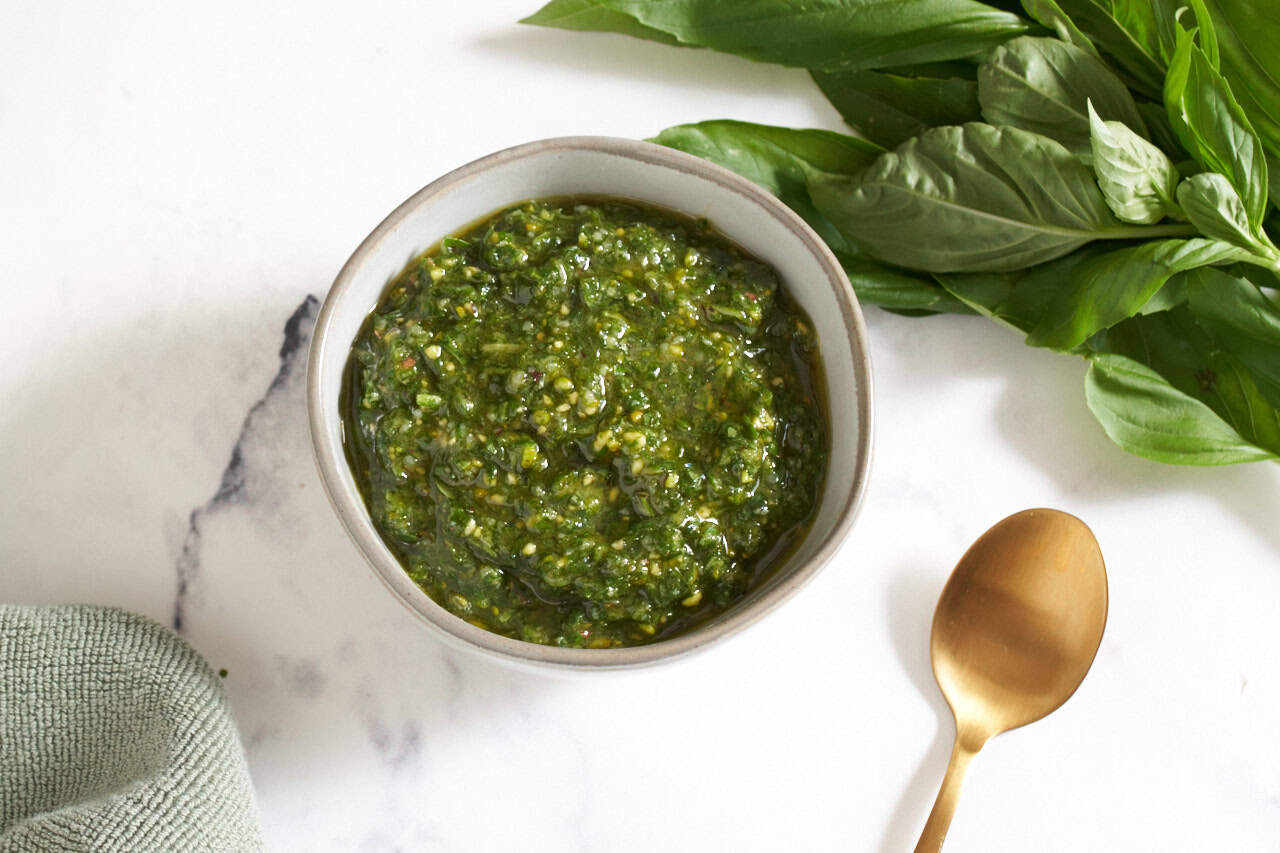A bowl of pistachio pesto, a gold spoon and fresh basil leaves are on the right.