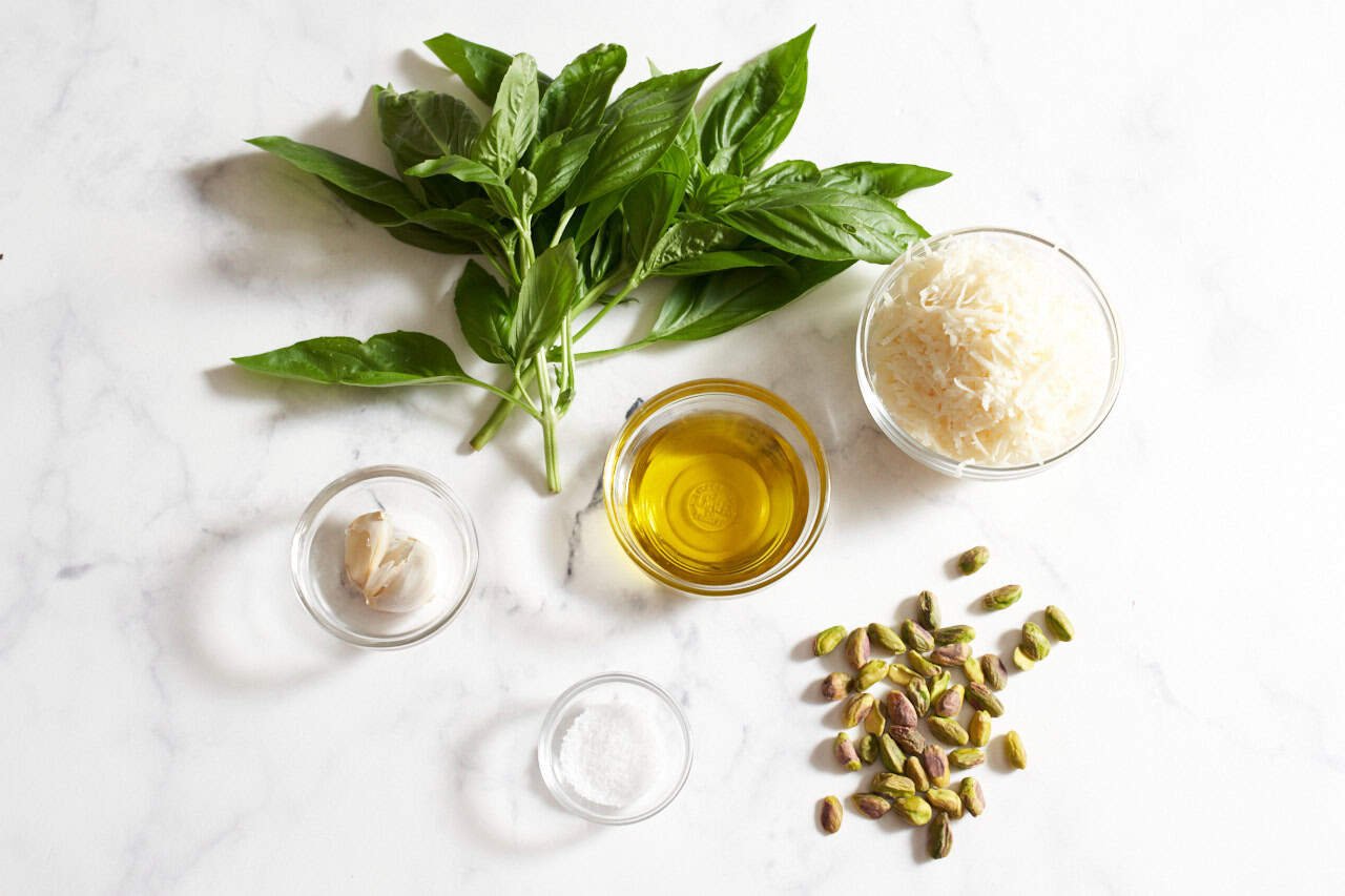 A bunch of fresh basil, some pistachios, and four small bowls of garlic cloves, grated parmesan cheese, olive oil, and kosher salt.