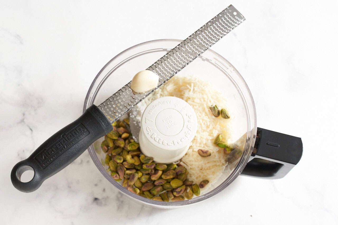 A garlic clove on a microplane grater positioned over a food processor full of pistachios and parmesan cheese.