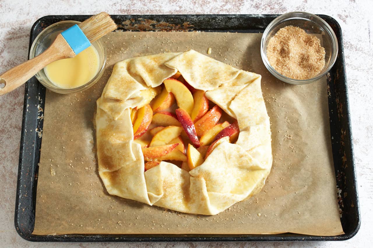 A puff pastry peach galette that has been brushed with egg wash and sprinkled with sugar, ready to go in the oven, bowls of egg wash and turbinado sugar in both the upper corners.
