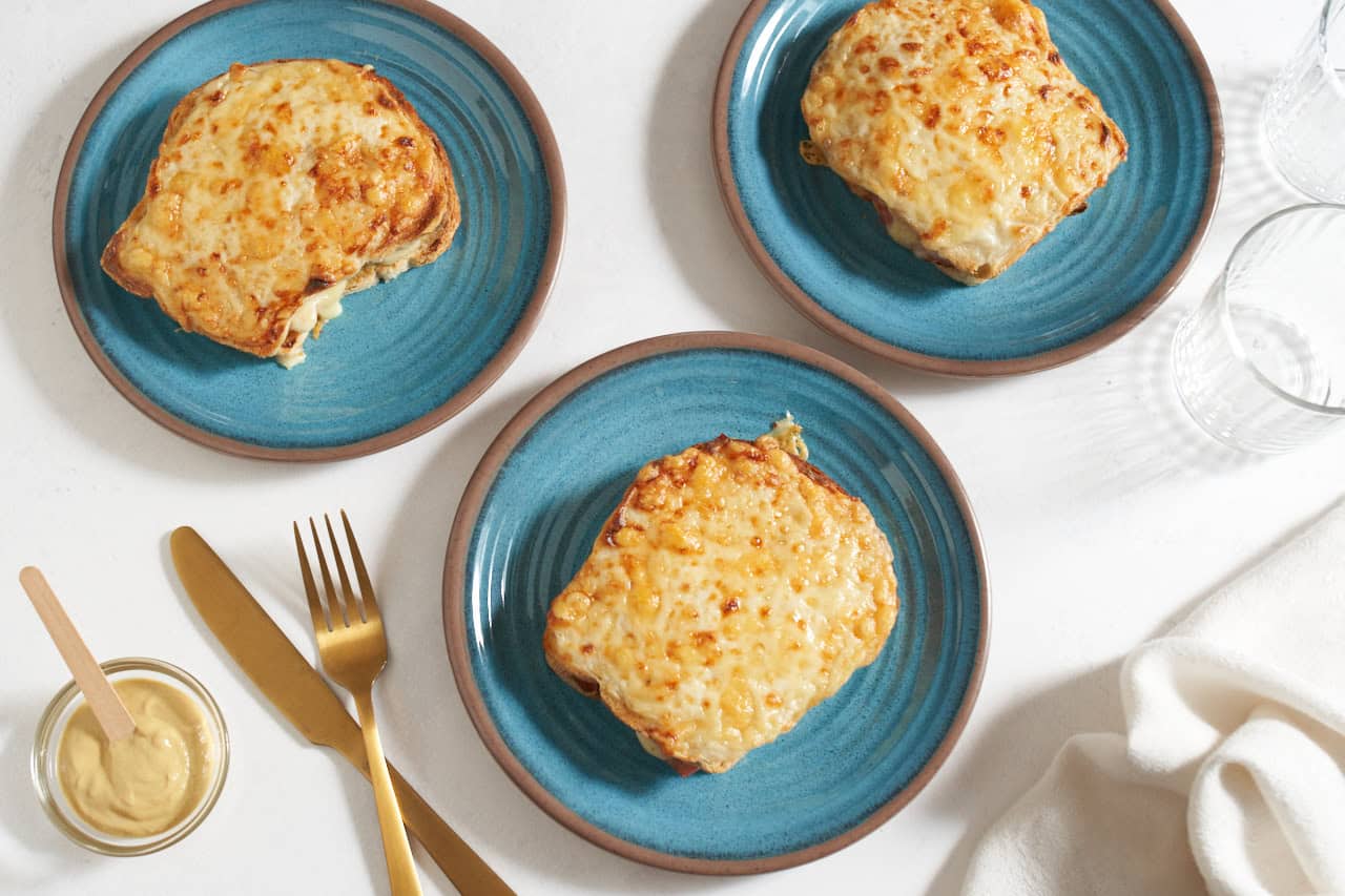 Three blue plates with one croque monsieur sandwich on each one, a gold knife and fork are on the left.