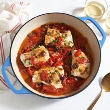Fish puttanesca in a skillet with blue handles, a gold spoon and two glasses of white wine are on the right.