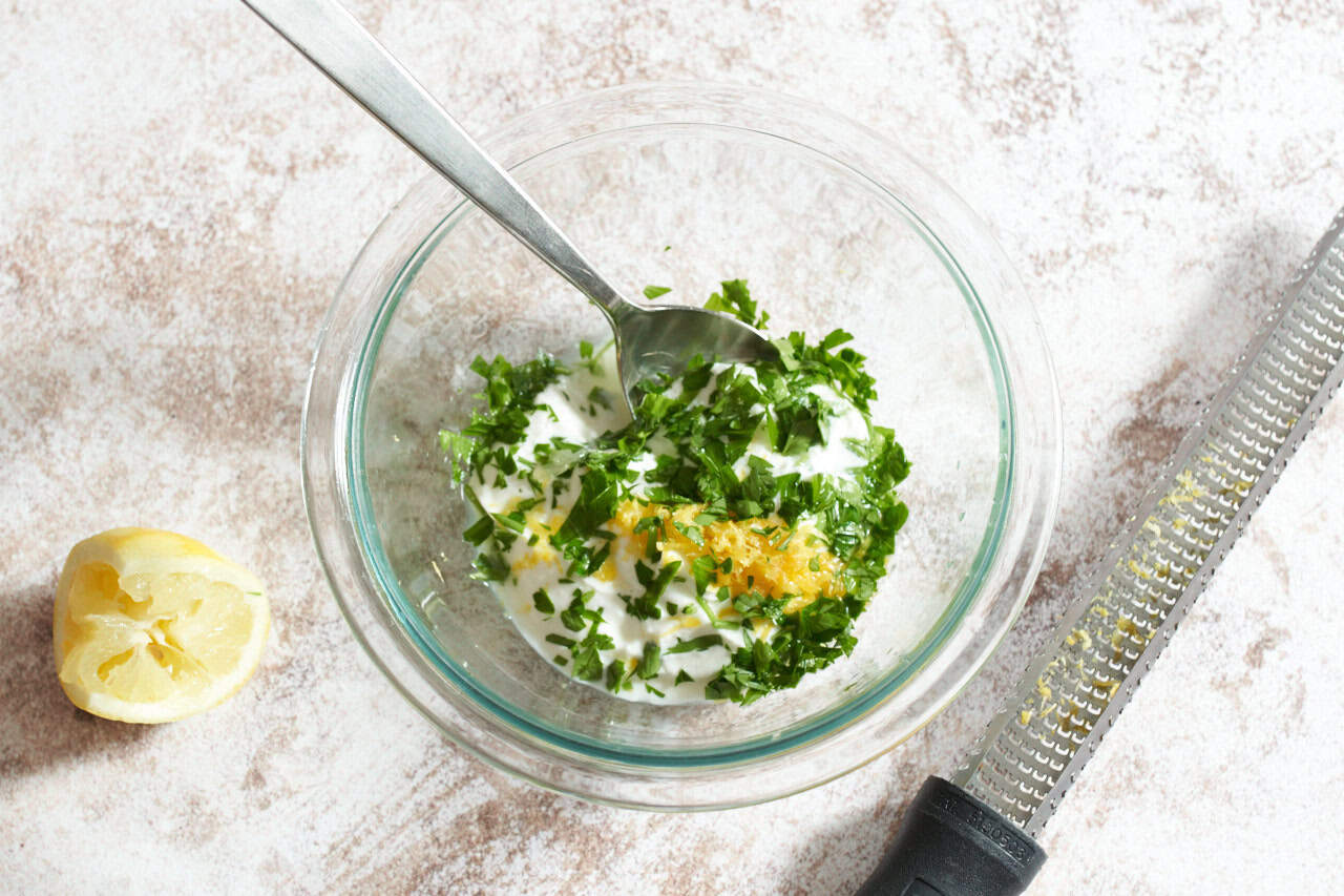 A bowl of yogurt topped with lemon zest and parsley, a grater is on the right, a squeezed lemon is on the left.