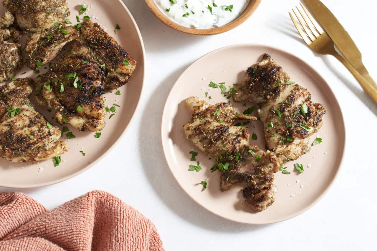 Grilled za'atar chicken on two pink plates, a bowl of yogurt sauce is above them, gold utensils are on the right.