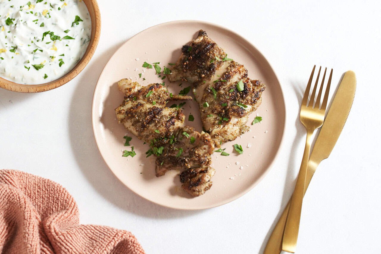 Grilled za'atar chicken on a pink plate, a bowl of yogurt sauce is on the left, gold utensils are on the right.