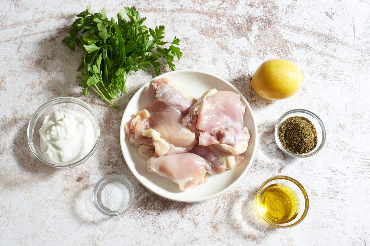 Chicken thighs on a white plate, a lemon, some fresh parsley, and small bowls of yogurt, olive oil, za'atar spice, and salt.