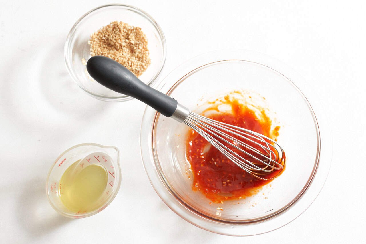 A whisk in a bowl of gochujang that has been mixed with sesame seeds and pickling liquid, a bowl of sesame seeds is in the upper right.