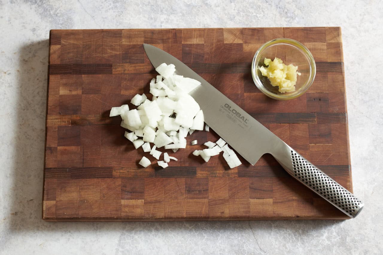A knife on a wooden cutting board with chopped onions and a small bowl of grated garlic.