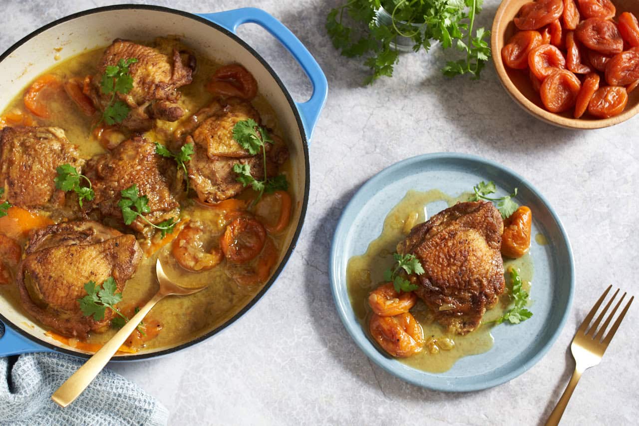 Chicken tagine with apricots in a blue skillet with a gold spoon in it, a plate with a piece of chicken and apricots is on the right, small bowls of cilantro and apricots are in the upper right.