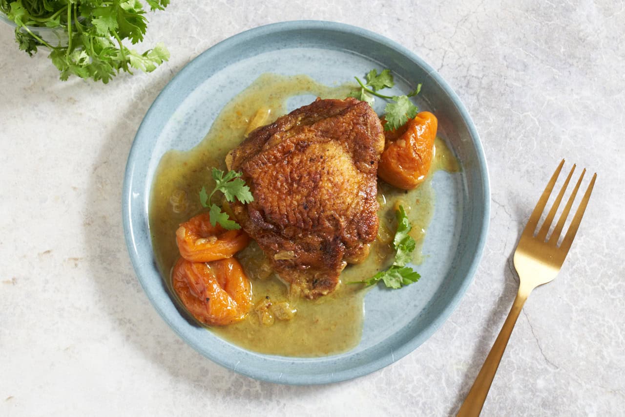 Chicken tagine with apricots and sauce garnished with cilantro on a blue plate, a gold fork is on the right.