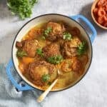 Chicken tagine with apricots garnished with fresh cilantro in a skillet with a gold spoon in it, a bowl of dried apricots is on the right, some fresh cilantro is in the upper left.