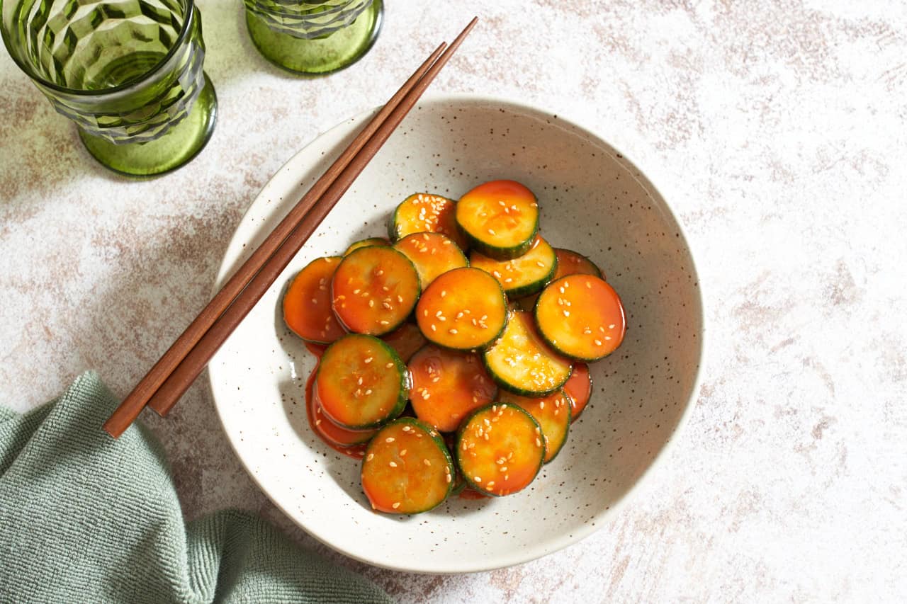 A bowl of spicy Asian cucumber salad topped with sesame seeds, chopsticks are resting on the left side of the bowl, two green glasses are in the upper left.