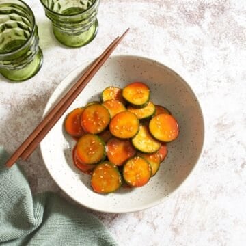 A bowl of spicy Asian cucumber salad topped with sesame seeds, chopsticks are resting on the left side of the bowl, two green glasses are in the upper left.