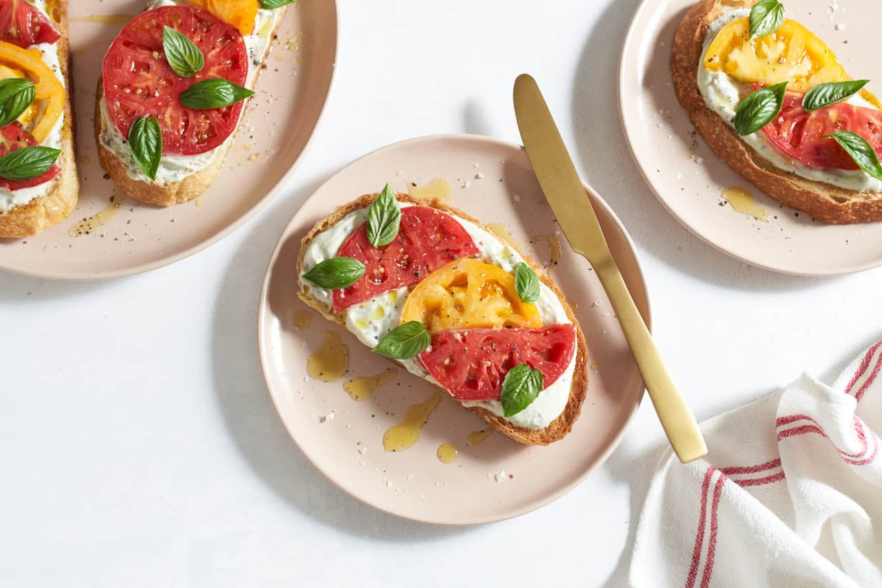 A tomato tartine topped with fresh basil and drizzled with olive oil on a pink plate with a gold knife, two more plates with tartines are in each upper corner.