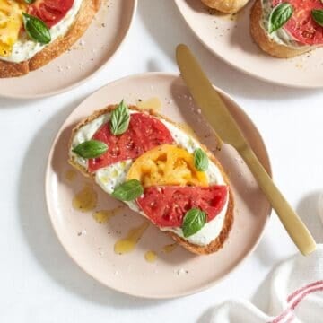 A tomato tartine topped with fresh basil and drizzled with olive oil on a pink plate with a gold knife, two more plates with tartines are in each upper corner.