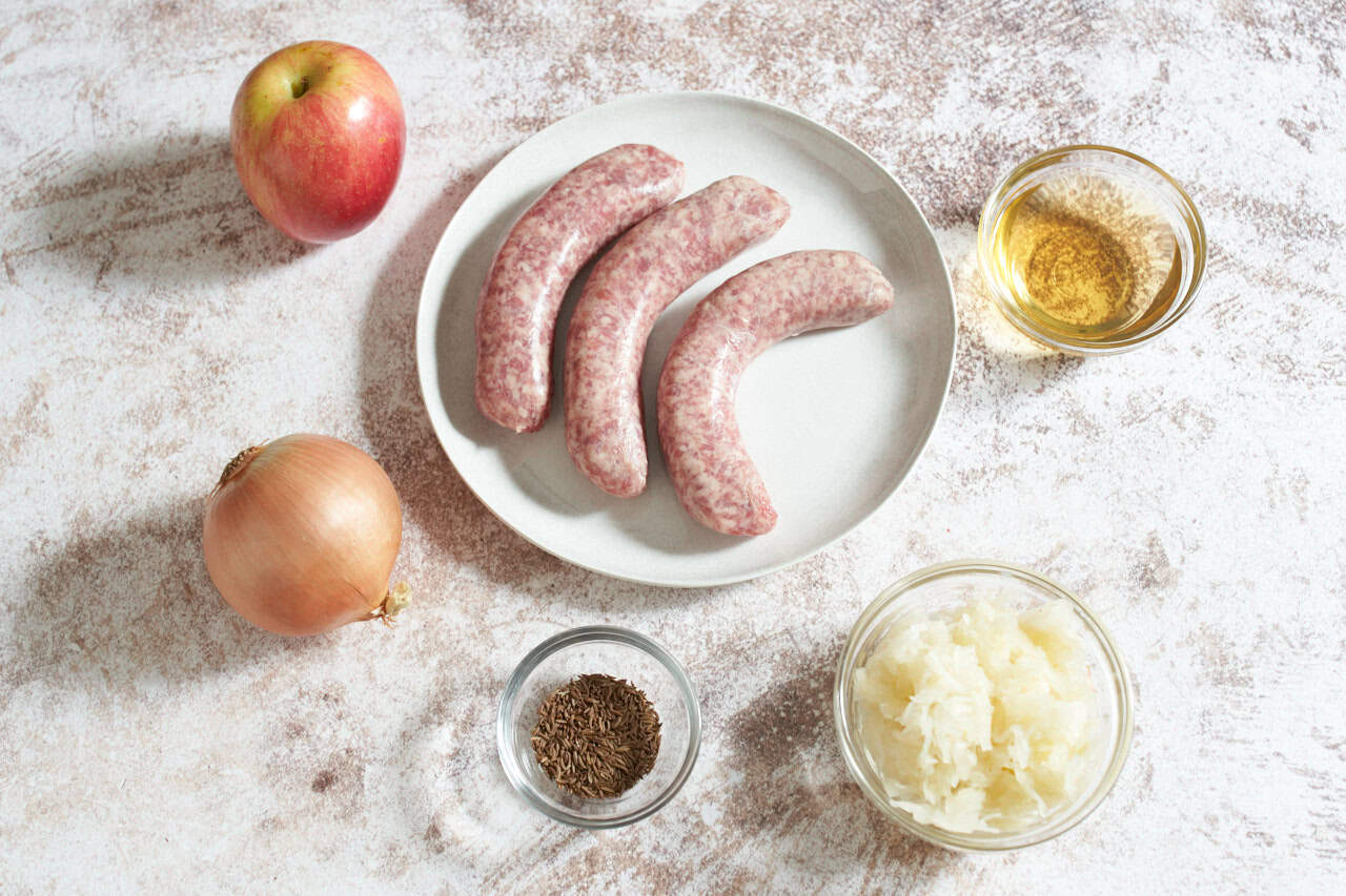 Three bratwurst on a white plate, an apple, an onion, and small bowls of beer, sauerkraut, and caraway seeds.