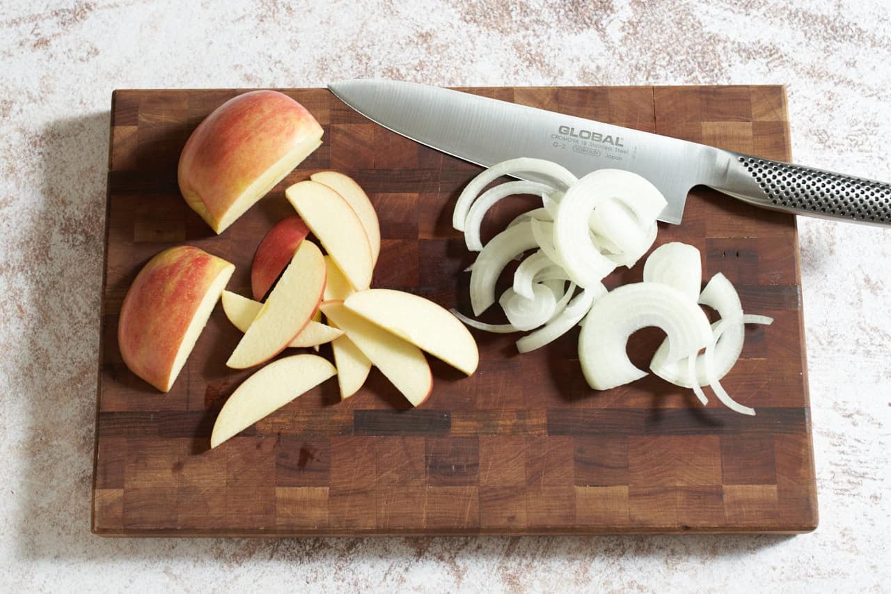 A knife on a cutting board with thinly sliced apples and onions.