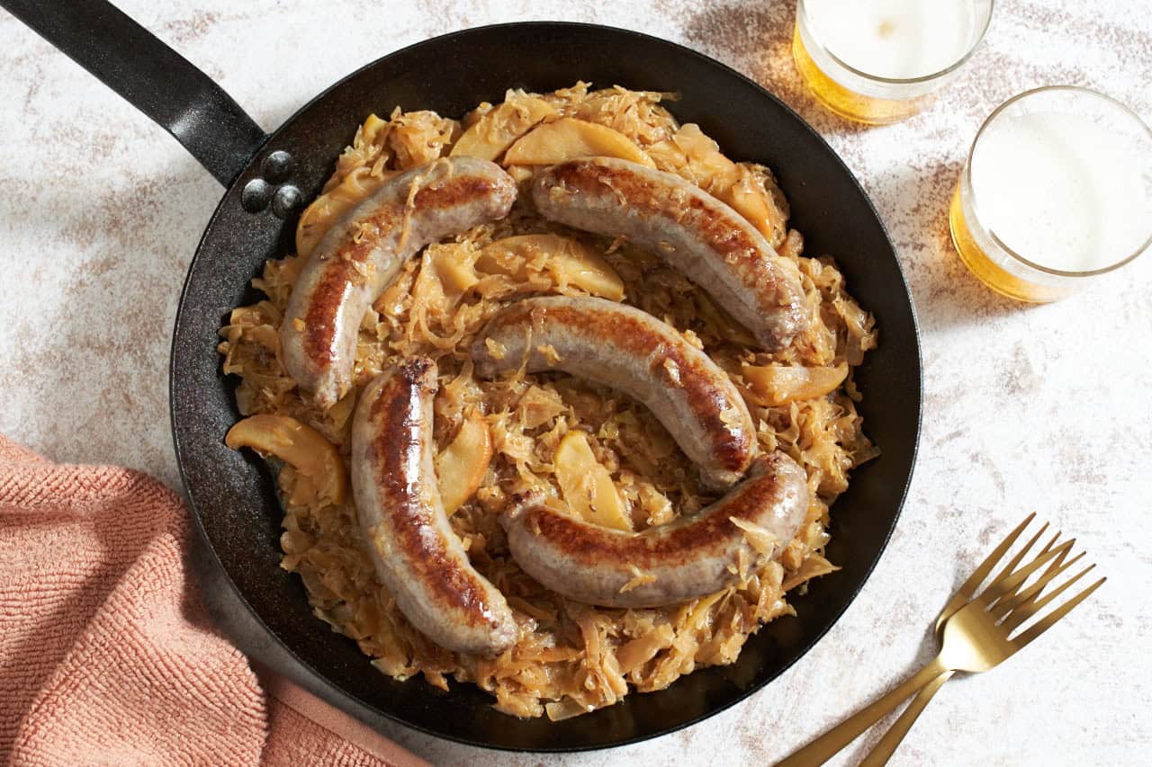 Bratwurst, sauerkraut, apples and onion in a black skillet, two glasses of beer and two gold forks are on the right.