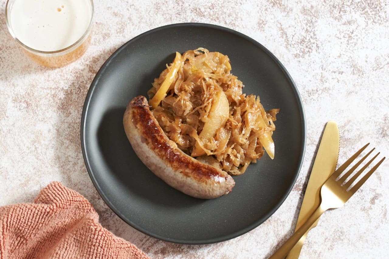 Bratwurst with sauerkraut on a black plate, a glass of beer is on the left, a gold knife and fork are on the right.