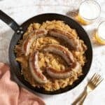 Bratwurst, sauerkraut, apples and onion in a black skillet, two glasses of beer and two gold forks are on the right.