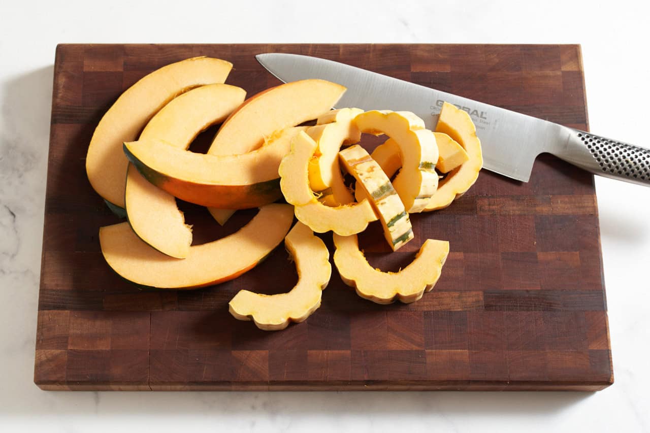 Sliced delicata and acorn squash on a wooden cutting board with a knife.