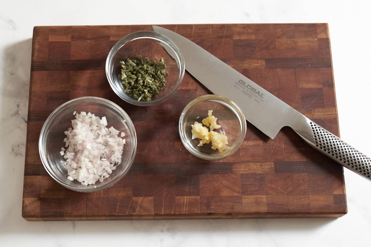 Small bowls of minced shallot, minced sage, and grated garlic on a wooden cutting board with a knife.