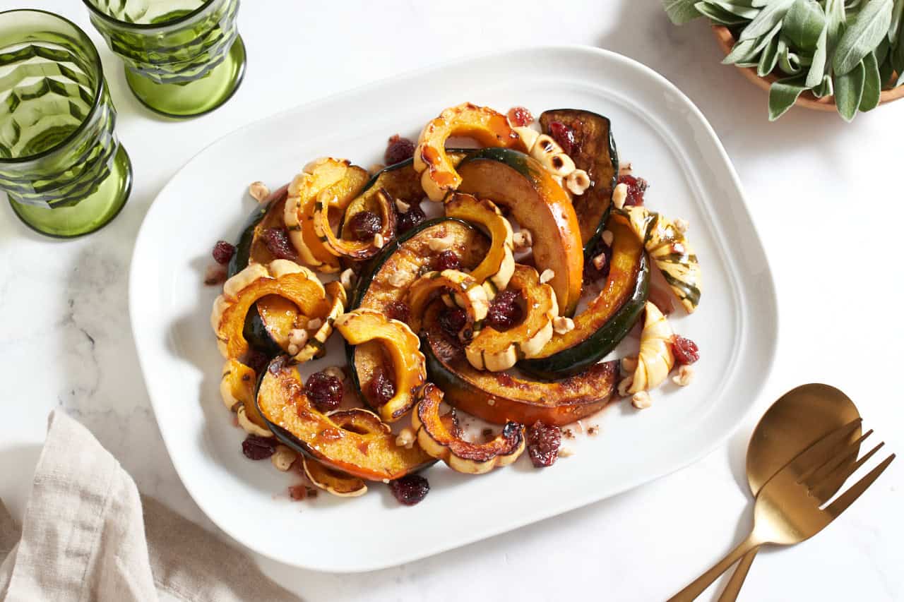 A platter of roasted winter squash topped with cranberry agrodolce sauce and hazelnuts, two green glasses are in the upper left, a bowl of fresh sage is in the upper right.