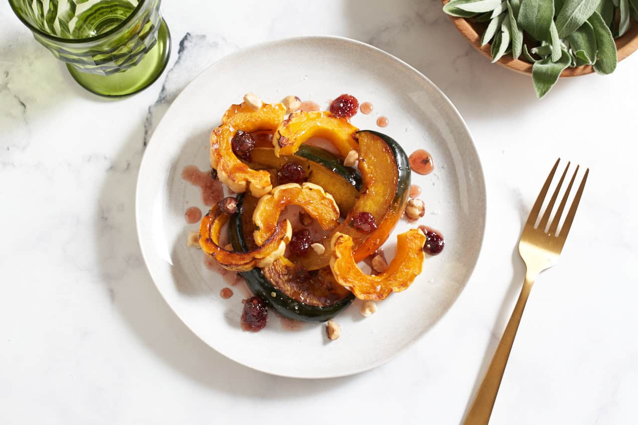 A plate of roasted winter squash topped with cranberry agrodolce sauce and hazelnuts, a green glass are in the upper left, a bowl of fresh sage is in the upper right, a gold fork is in the lower right.