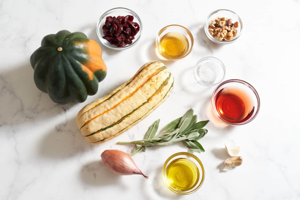 An acorn squash and a delicata squash, some sprigs of fresh sage, a shallot, a garlic clove, and small bowls of olive oil, red wine vinegar, honey, dried cranberries, and hazelnuts.