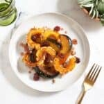 A plate of roasted winter squash topped with cranberry agrodolce sauce and hazelnuts, a green glass are in the upper left, a bowl of fresh sage is in the upper right, a gold fork is in the lower right.