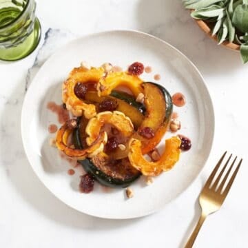 A plate of roasted winter squash topped with cranberry agrodolce sauce and hazelnuts, a green glass are in the upper left, a bowl of fresh sage is in the upper right, a gold fork is in the lower right.